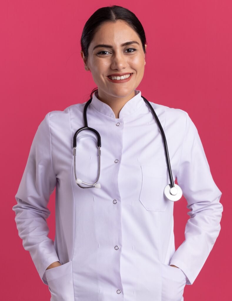young woman doctor in medical coat with stethoscope looking at camera smiling with happy face standing over pink background